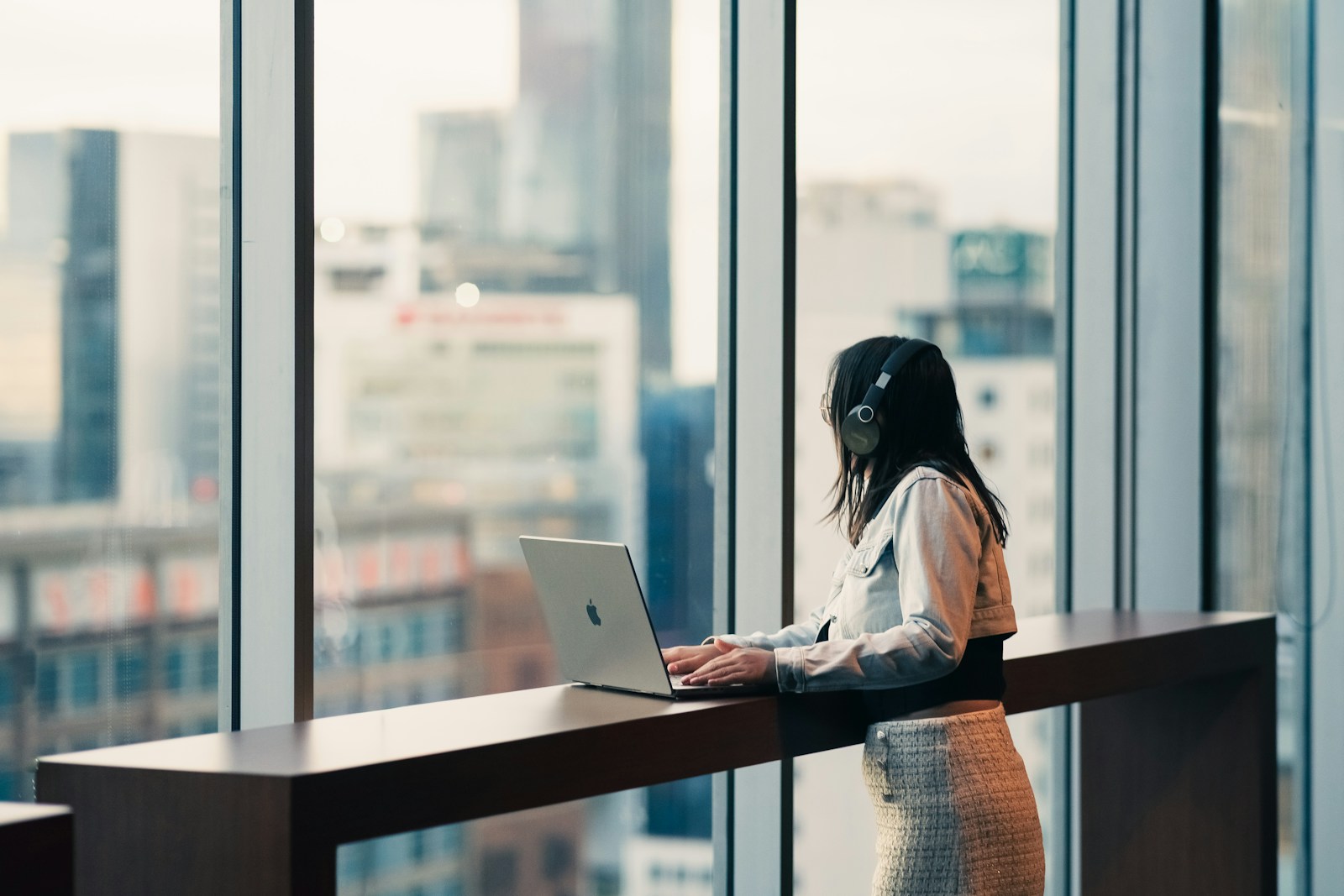 Woman wearing headphones works on laptop by window.